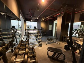 Une salle de sport équipée de divers appareils d’exercice, d’un banc et offrant une vue sur la ville grâce à de grandes fenêtres.