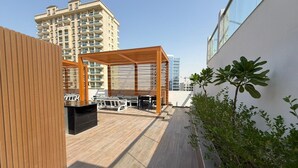 A rooftop terrace with a wooden pergola, outdoor seating, and a view of high-rise buildings. - Bright and Modern One Bedroom Apartment (Arjan-Dubailand)