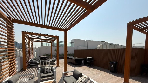 An outdoor patio area with wooden pergolas, multiple seating arrangements, and a barbecue grill. - Bright and Modern One Bedroom Apartment (Arjan-Dubailand)