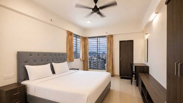 A hotel room with a bed, a desk, a chair, a wardrobe, and a ceiling fan. - Deluxe Hotel Urban Oasis (Pune)