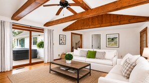 A spacious living room with a high ceiling, wooden beams, a white sofa, a coffee table, and a sliding glass door leading to an outdoor area.