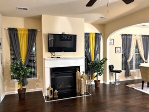 A living room with a fireplace, a television mounted above it, and a dining area with a chair and table.