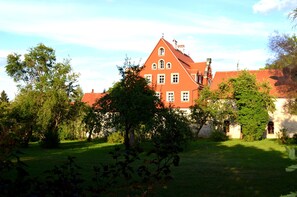A large, red-roofed building with multiple windows, surrounded by lush greenery and trees. - Our "Loft" vacation suite in the "Schlossberg" vacation home (Betzenstein)