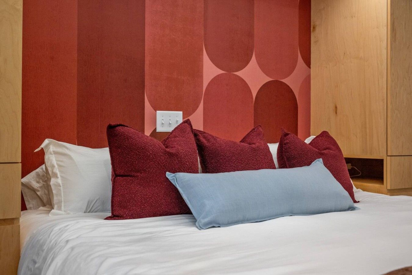 A bed with white bedding and multiple pillows, including a red and a blue one, in a room with wooden furniture and a red wall with a geometric pattern.
