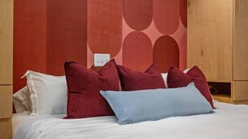 A bed with white bedding and multiple pillows, including a red and a blue one, in a room with wooden furniture and a red wall with a geometric pattern.