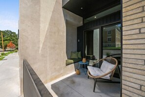 A modern outdoor seating area with a wicker chair and a small round table, a cushioned bench, and a potted plant. - Designer 1BR at City Park | Walk to Aggieville (Manhattan)