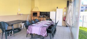 A covered outdoor dining area with a long table set for multiple people, chairs, a small kitchen area with a brick oven, and a refrigerator. - Maison avec piscine privée (Amor)