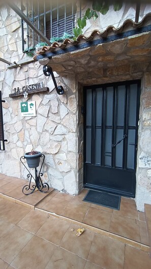 Un bâtiment rustique en pierre, avec une porte noire, une enseigne indiquant « CASAS RURALES » et un pot de fleurs posé sur le sol.
