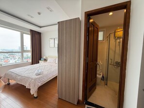 Quarto de hotel com cama, janela com cortinas, porta de madeira para um banheiro com chuveiro e vista para a cidade.