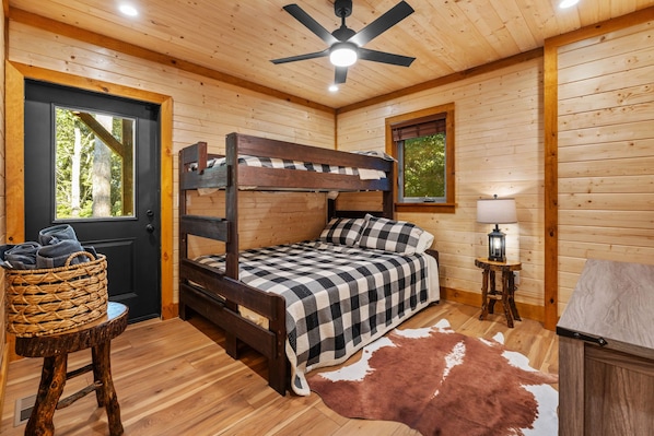A wooden cabin bedroom with a bunk bed, a single bed, a lamp, a rug, and a door with a view of trees.