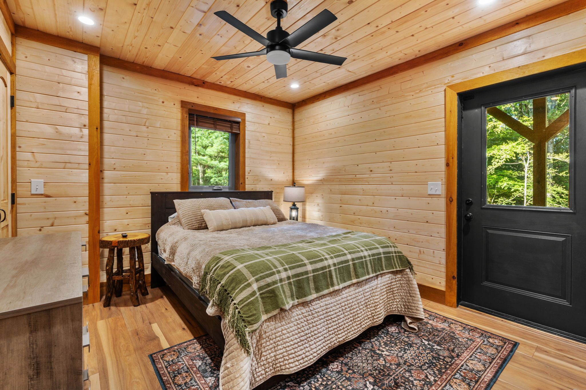 Un dormitorio de cabaña de madera con cama, mesita de noche, lámpara, ventana y puerta.
