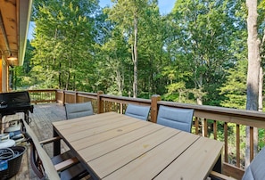 An outdoor deck with a wooden table and chairs, surrounded by a wooden railing, overlooking a forested area.
