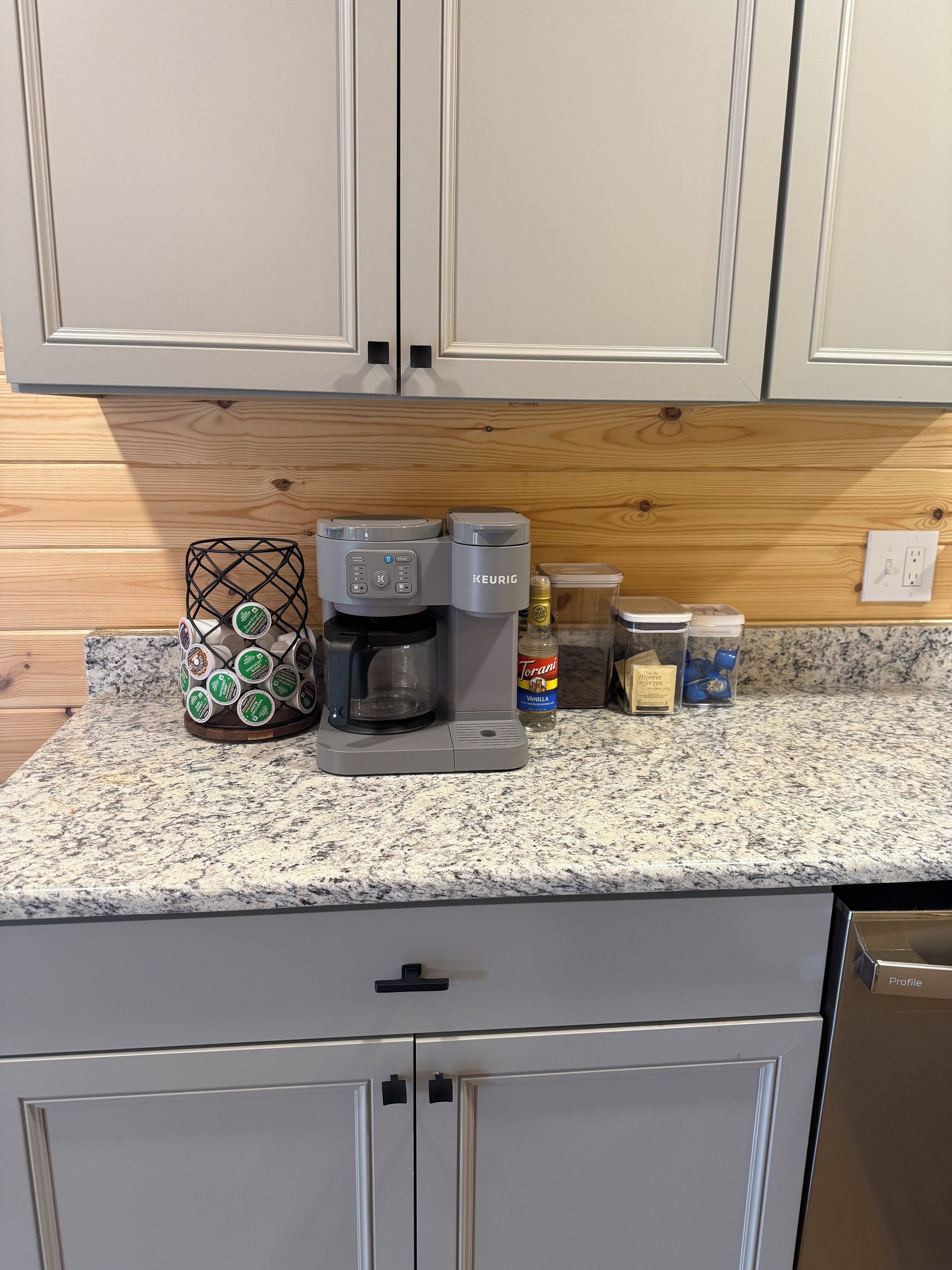 Een keuken met een koffiezetapparaat, een houder voor koffiekapsules, een fles Torani-siroop en een verpakking koffiekapsules.