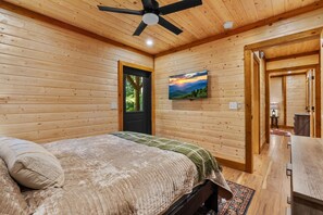 Une chambre à coucher comprenant un lit, un ventilateur de plafond, un téléviseur fixé au mur et une porte donnant sur une autre pièce.