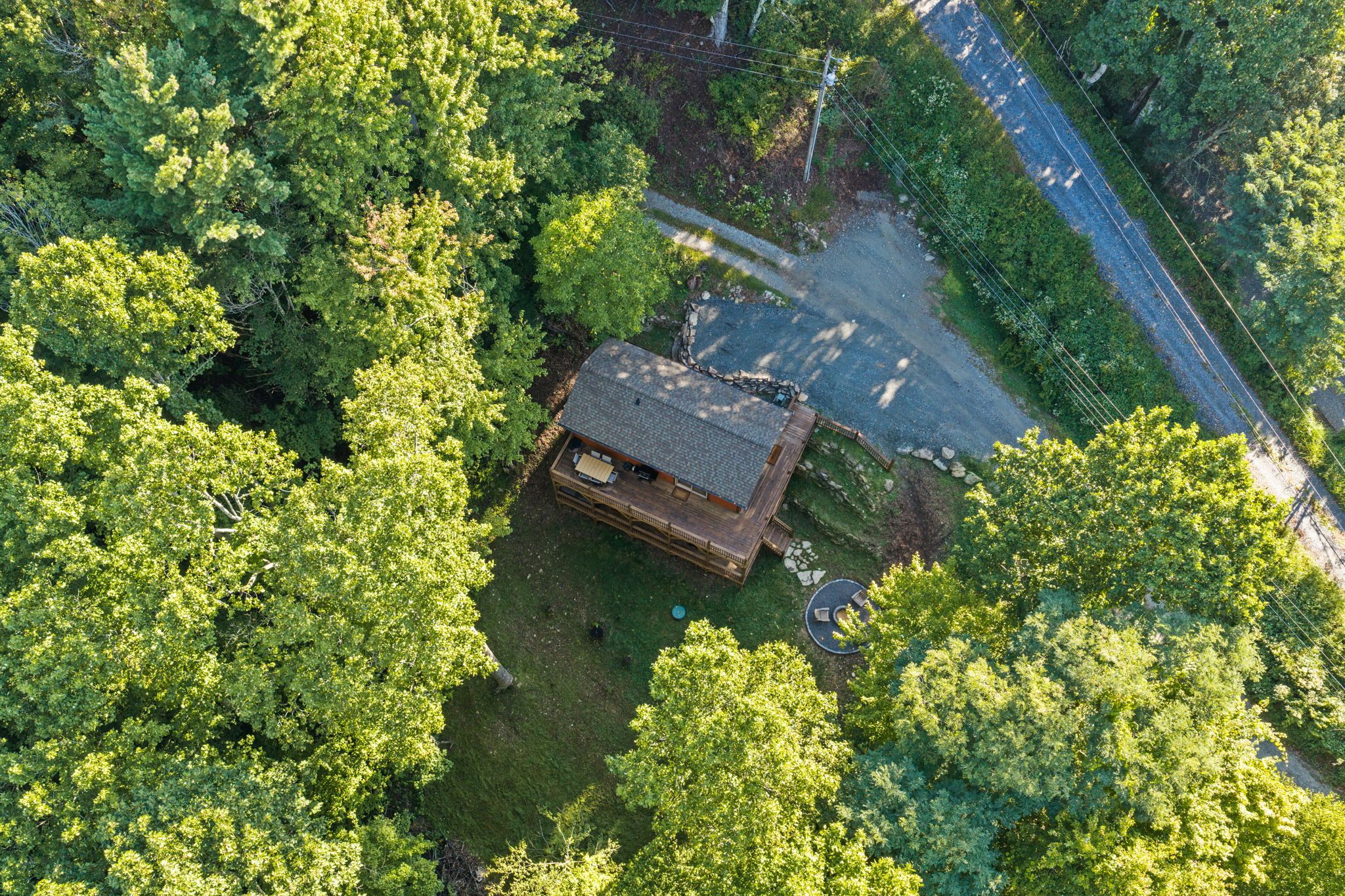 An aerial view of a cabin surrounded by dense forest, with a gravel driveway and a wooden deck.