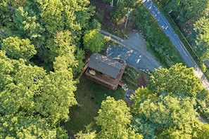 An aerial view of a cabin surrounded by dense forest, with a gravel driveway and a wooden deck.