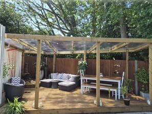 Ein überdachter Patio-Bereich mit Holztisch und Sitzgelegenheiten, umgeben von Pflanzen und einem Holzzaun.