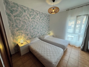 Ein Zimmer mit zwei Betten, einem gemusterten Wandpapier, einer Hängelampe und einem Fenster mit Vorhängen.