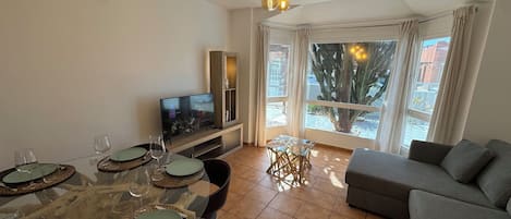 Una sala moderna con un sofá gris, una mesa de comedor con platos y vasos, un televisor sobre un mueble de madera, y un ventanal con vistas a una palmera y edificios.
