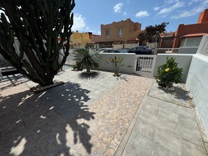 A courtyard with a cactus, a palm tree, and a small bush. There are cars parked in the background and a building with a beige facade. - The Ocean Sur Beach House: A Villa with Ocean Views (Corralejo)