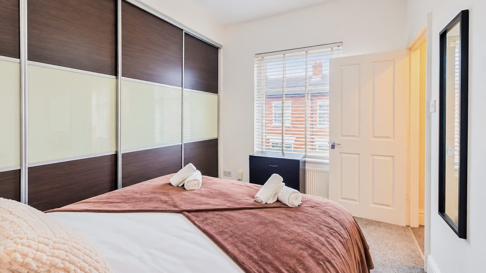 A bedroom with a bed, a wardrobe, a bedside table, and a window with blinds.
