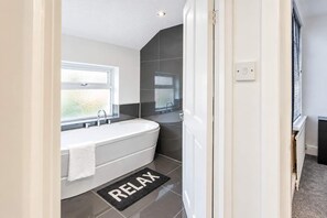 A modern bathroom with a freestanding bathtub, a window, and a 'RELAX' mat. - Charming 3BR Home | Pool Table | Free Parking (Manchester)