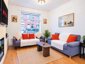 A living room with two sofas, a coffee table, a TV, and a large window with a view of a brick building. - Charming 3BR Home | Pool Table | Free Parking (Manchester)