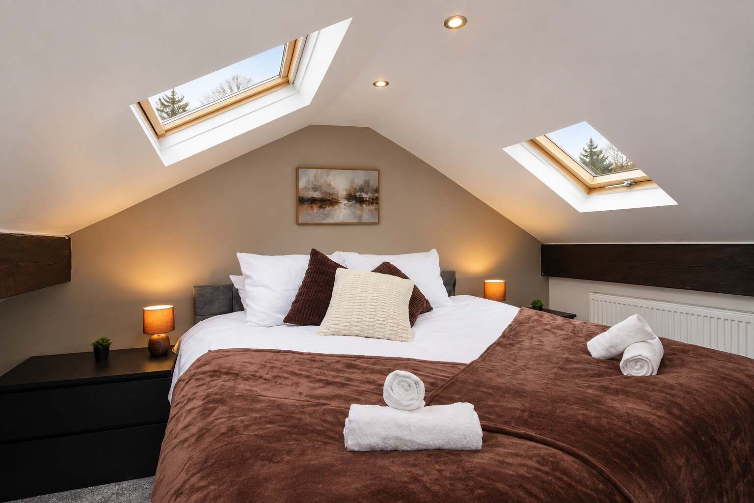 Un dormitorio con techo inclinado, tragaluces, una cama con manta marrón, almohadas blancas y un cuadro en la pared.