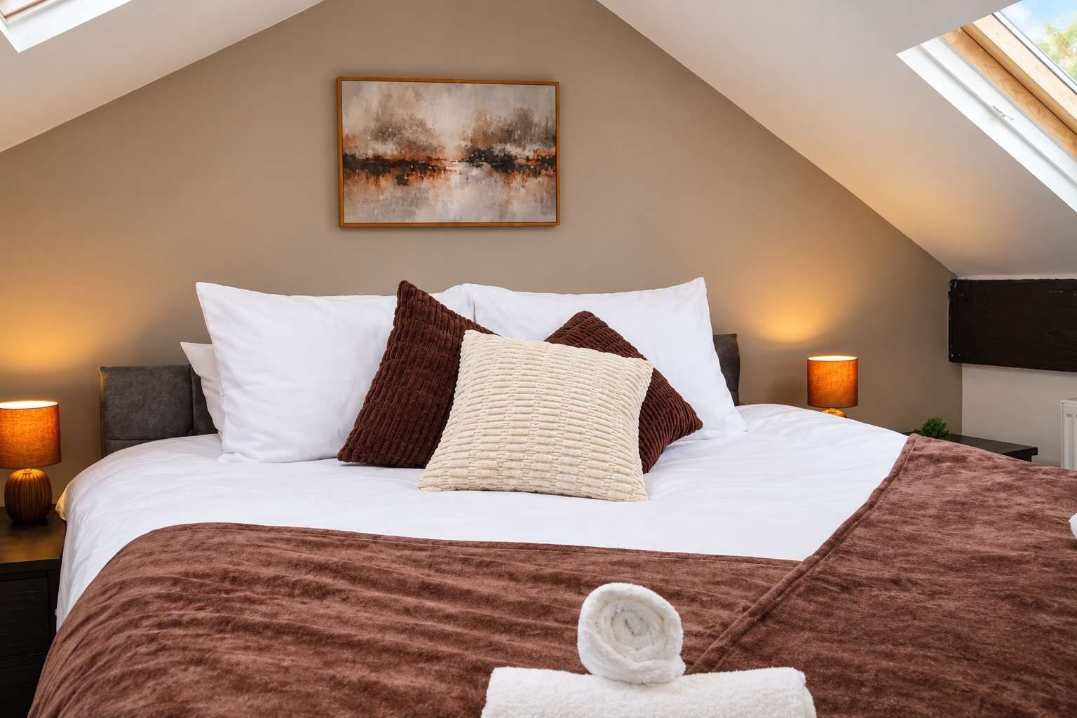 A neatly made bed with a brown blanket and pillows, a painting on the wall, and a window providing natural light.