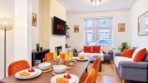 Un salon moderne comprenant un coin repas, un téléviseur à écran plat et une vue sur un immeuble grâce à de grandes fenêtres.