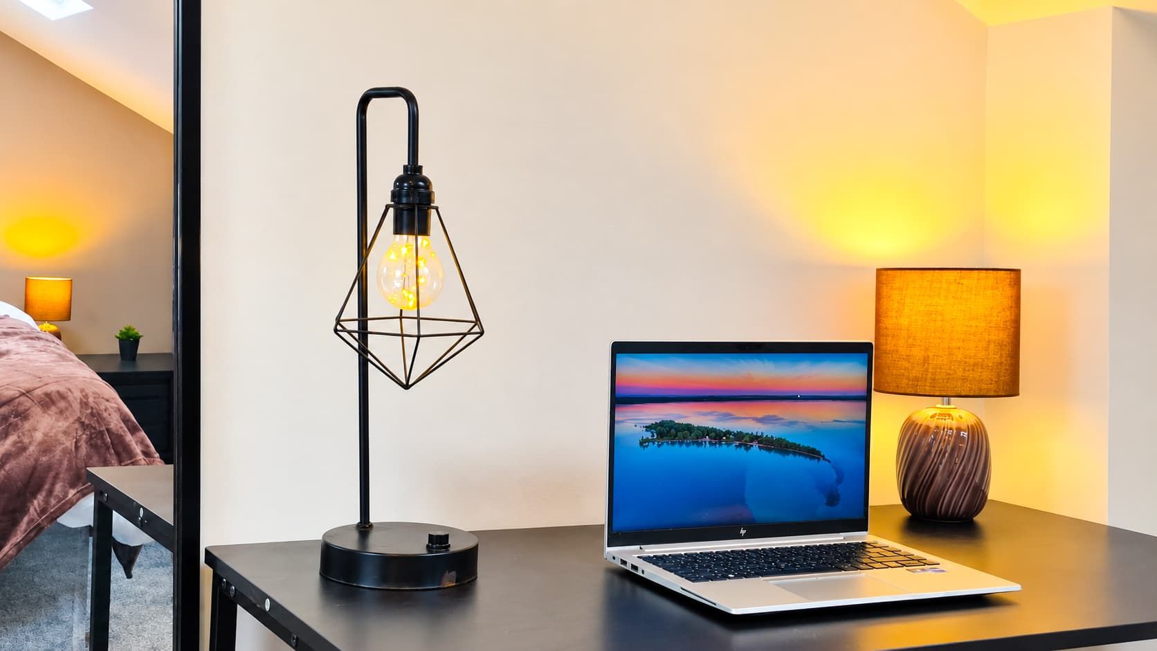A laptop on a desk with a geometric lamp, a laptop stand, and a lamp with a textured lampshade.