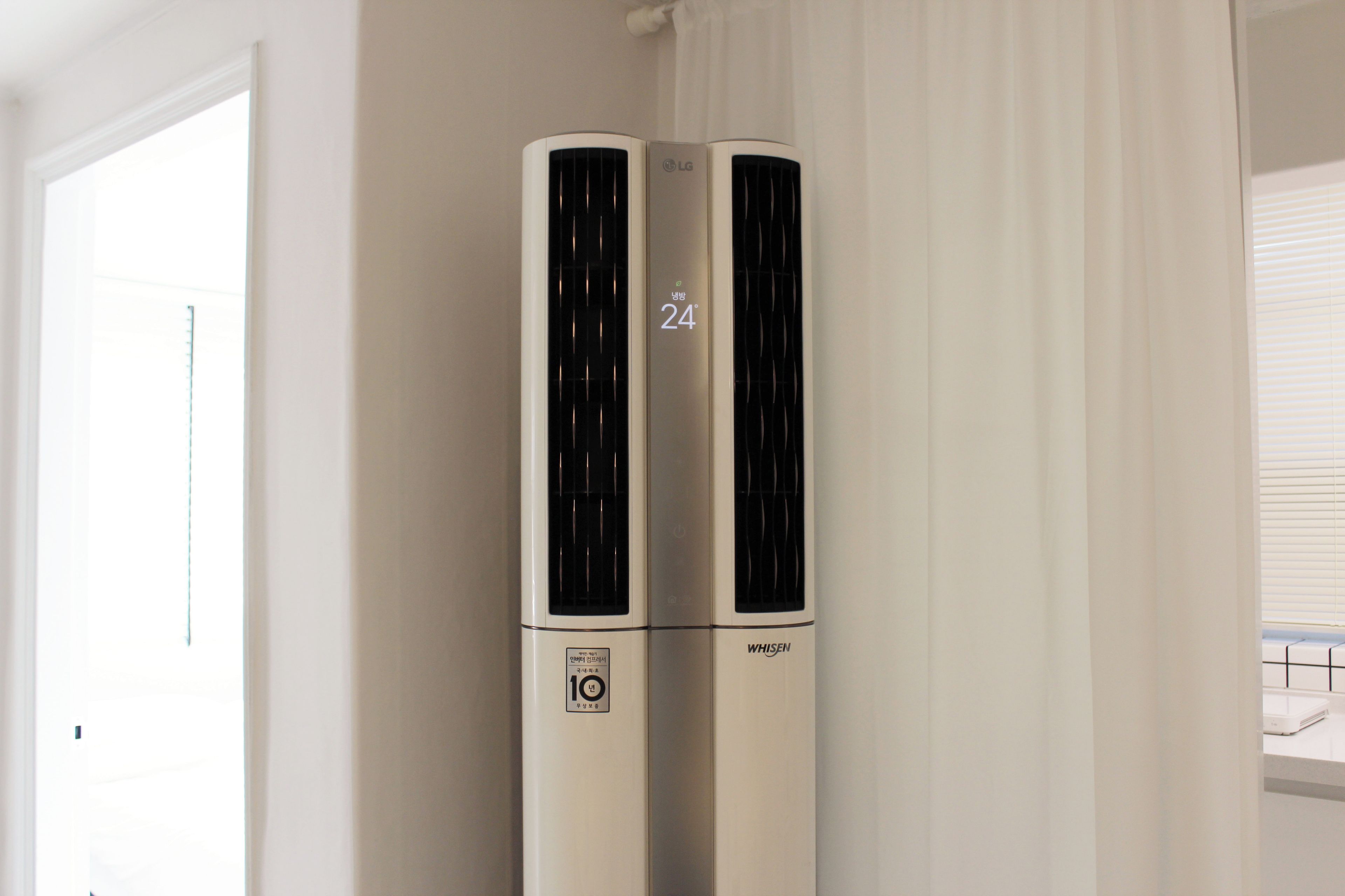 A white air conditioning unit with a digital display indicating 24 degrees Celsius, placed in a room with white walls and a window with blinds.