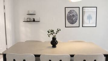 A dining area with a long table, four chairs, a vase with plants, and two framed artworks on the wall.