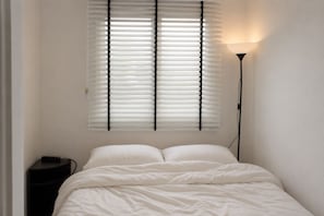 Ein Schlafzimmer mit einem Bett, einem Nachttisch, einer Lampe und einem Fenster mit Jalousien.