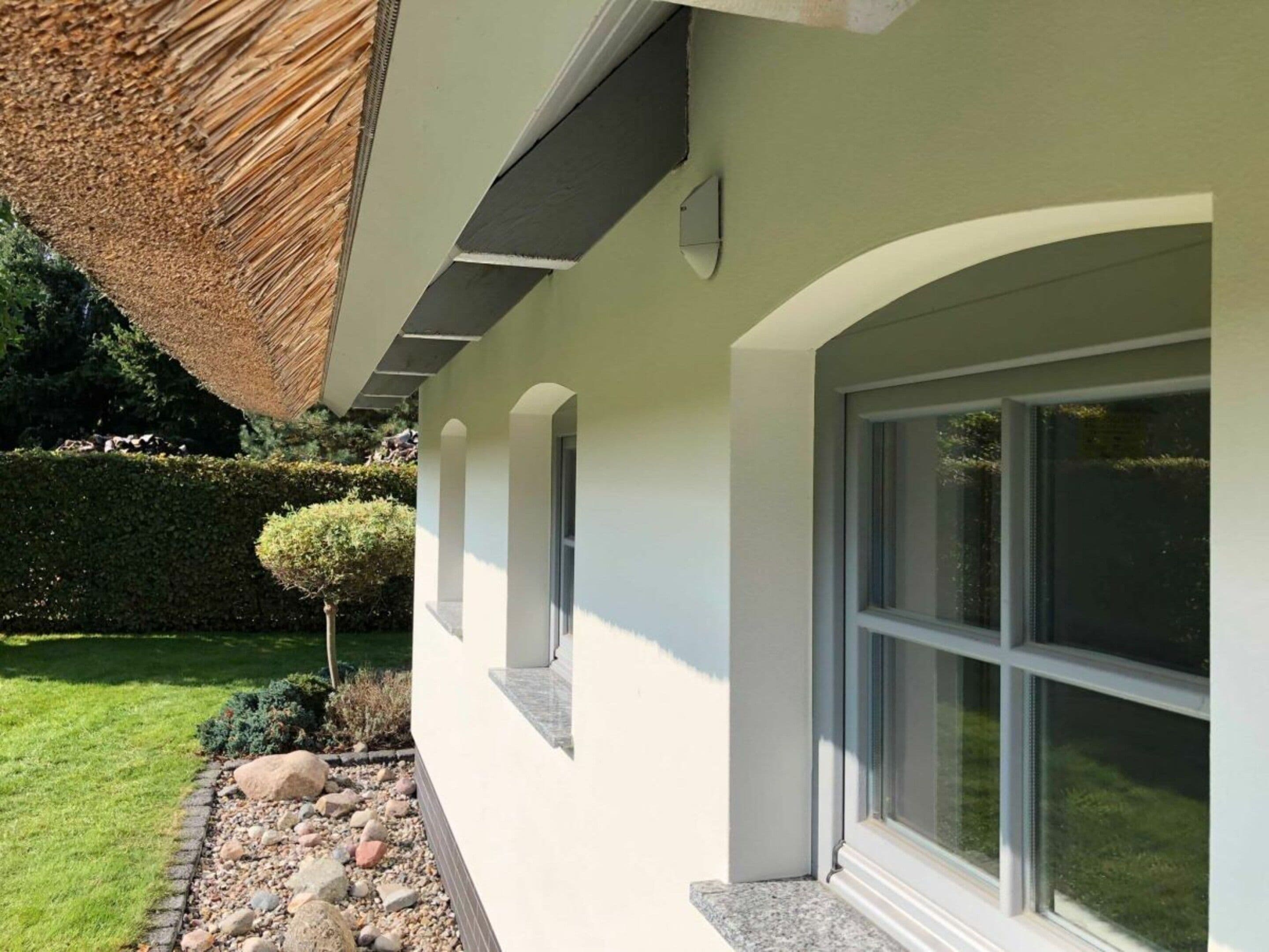 Un bâtiment moderne doté d’une grande fenêtre, d’un petit arbre et d’un jardin bien entretenu, agrémenté d’une haie et de rochers.