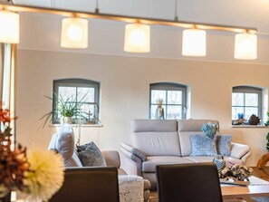 Ein Wohnzimmer mit einer Couch, Sesseln und einem Couchtisch. Es gibt Fenster mit Vorhängen, eine Pflanze und eine Vase auf der Fensterbank.