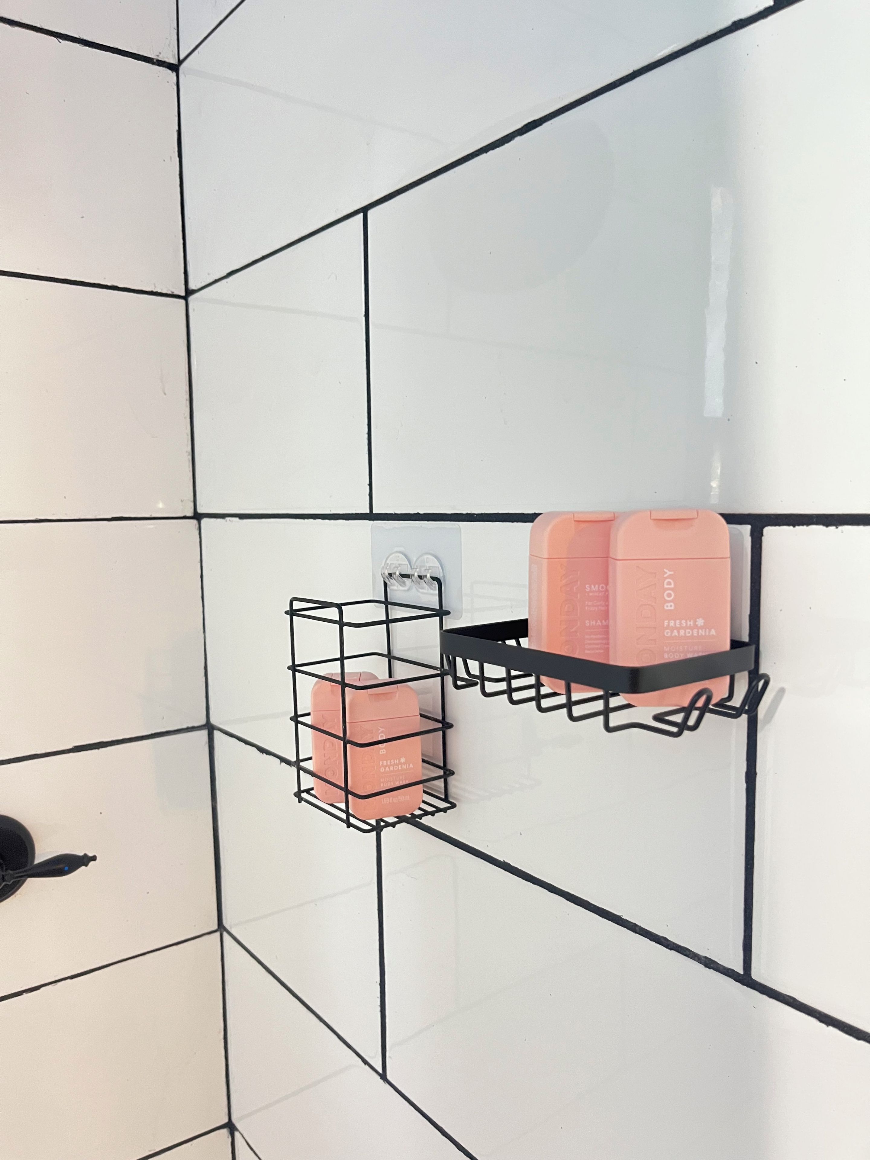 Un baño con paredes de azulejos blancos y un estante metálico negro que contiene una botella rosa y un recipiente transparente.