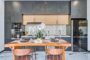 A modern kitchen with dark cabinets, a wooden dining table set for two, and a central island with a vase of white flowers. - Stylish 3BR Pool Villa in Canggu, Bali (Bali)