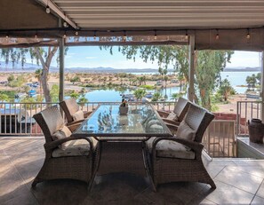 An outdoor dining area with a glass table and wicker chairs, overlooking a body of water and distant mountains. - Lakefront Home — Marina & Sunset Views! (Lake Havasu City)