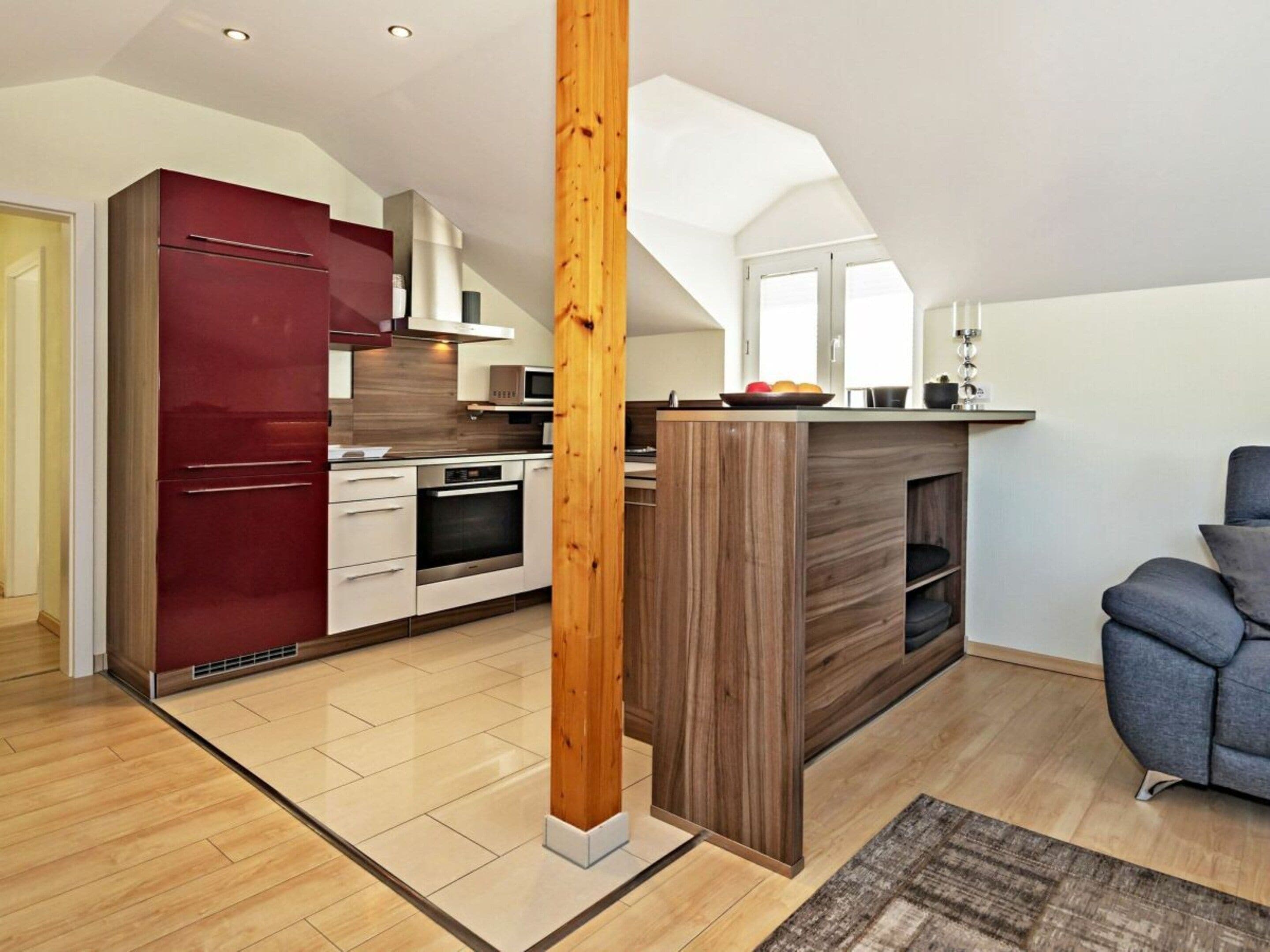 A modern kitchen with a wooden column, a red refrigerator, and a gray sofa.