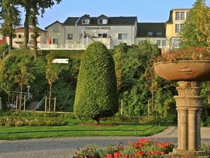 A landscaped garden with a well-maintained hedge, a stone fountain, and a building with a sign that reads 'TERBASSEN-KAFFEE' in the background. - Holiday Apartment With 1 Bedroom (Heringsdorf)