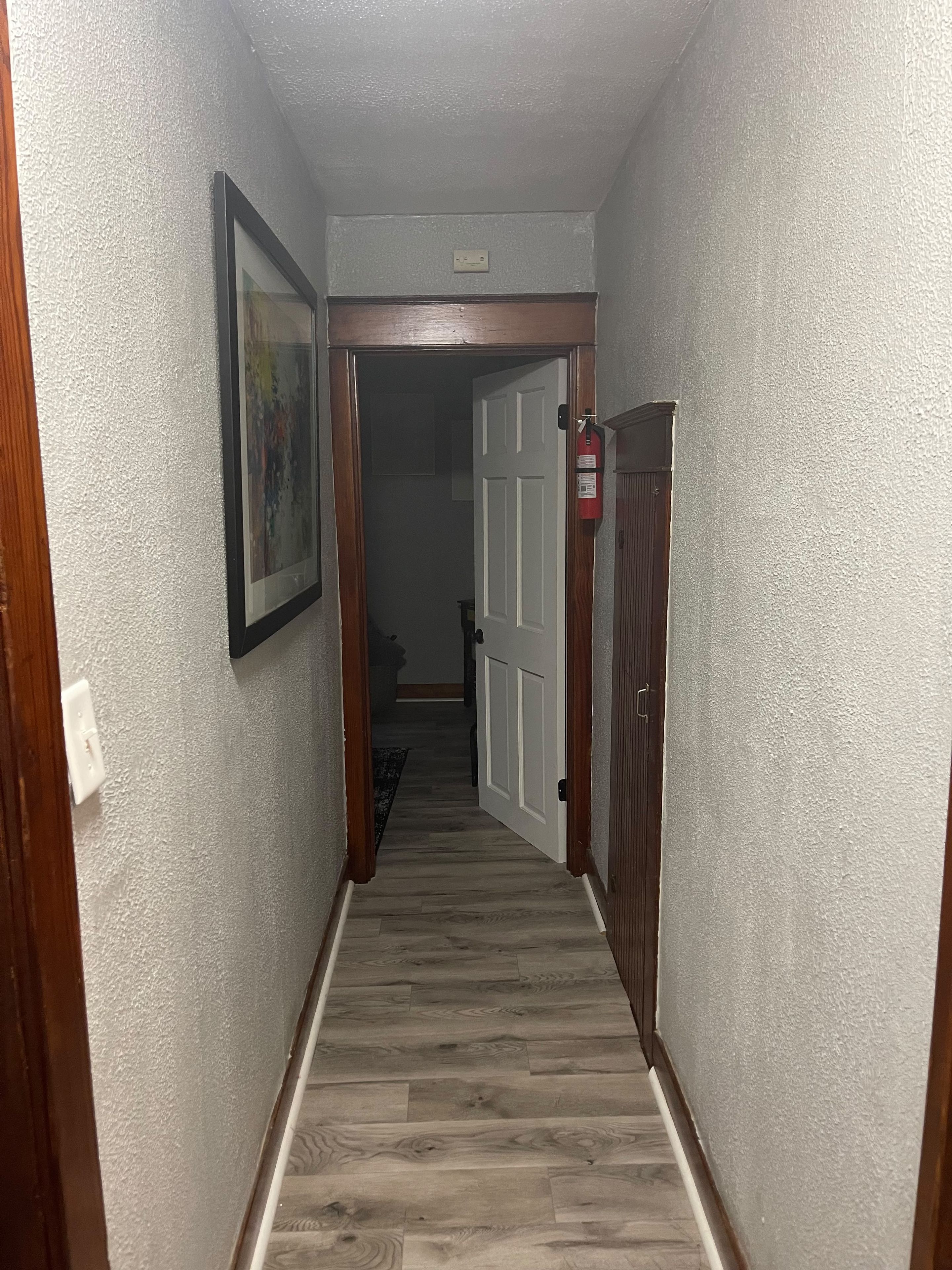 Un couloir avec une porte en bois, un extincteur et un tableau encadré accroché au mur.