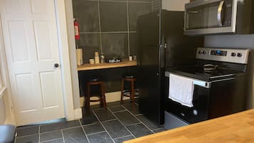 A compact kitchen with a black refrigerator, a microwave, and a stove. There are two stools and a small shelf with kitchen items.