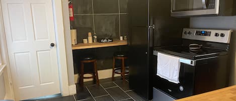 Une cuisine compacte avec un réfrigérateur noir, un four à micro-ondes et une cuisinière. On y trouve deux tabourets et une petite étagère contenant des ustensiles de cuisine.