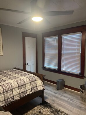 Une chambre à coucher avec un lit, un ventilateur de plafond, une fenêtre avec des stores, une porte et un petit tapis.