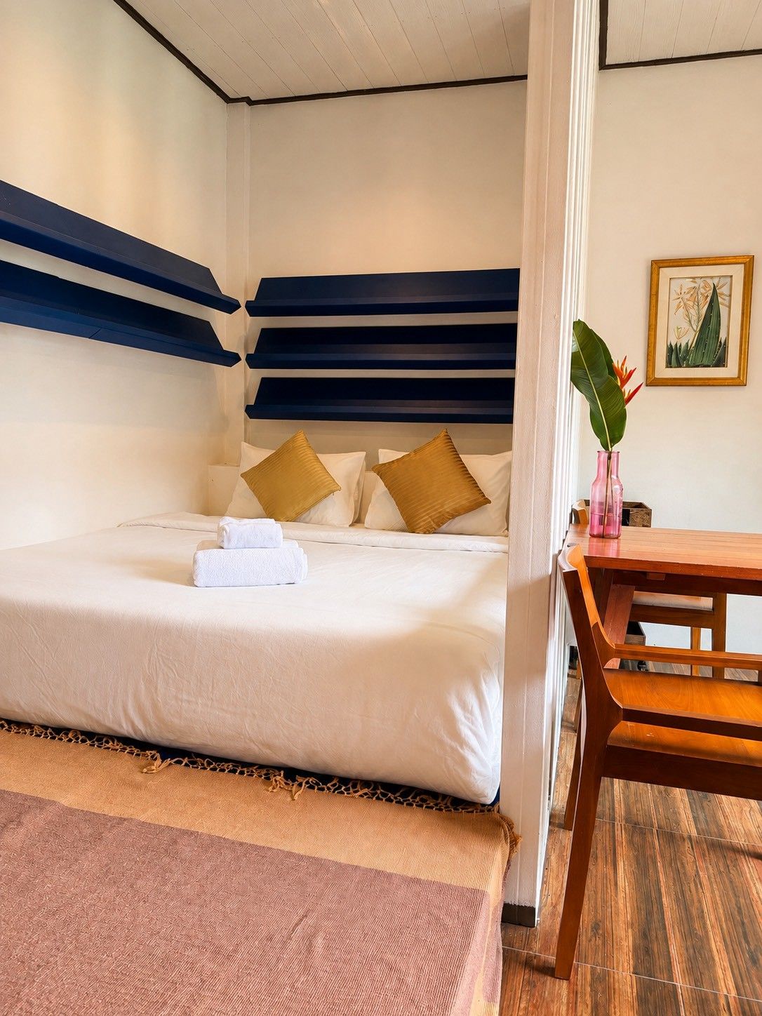 Una cama bien hecha con sábanas blancas y almohadas doradas, una mesa de madera con una silla, e impresión botánica enmarcada en la pared.