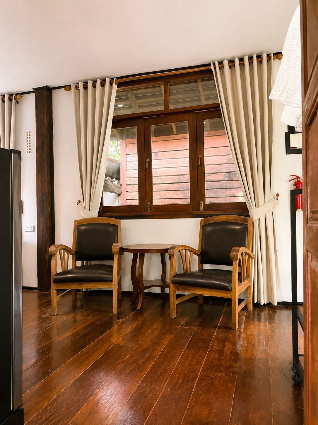 A room with two wooden chairs, a small table, a window with curtains, and a fire extinguisher on the wall.