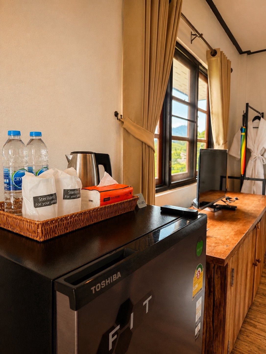 A room with a Toshiba refrigerator, a tray with bottled water and sterilized bags, a coffee pot, and a window with curtains.