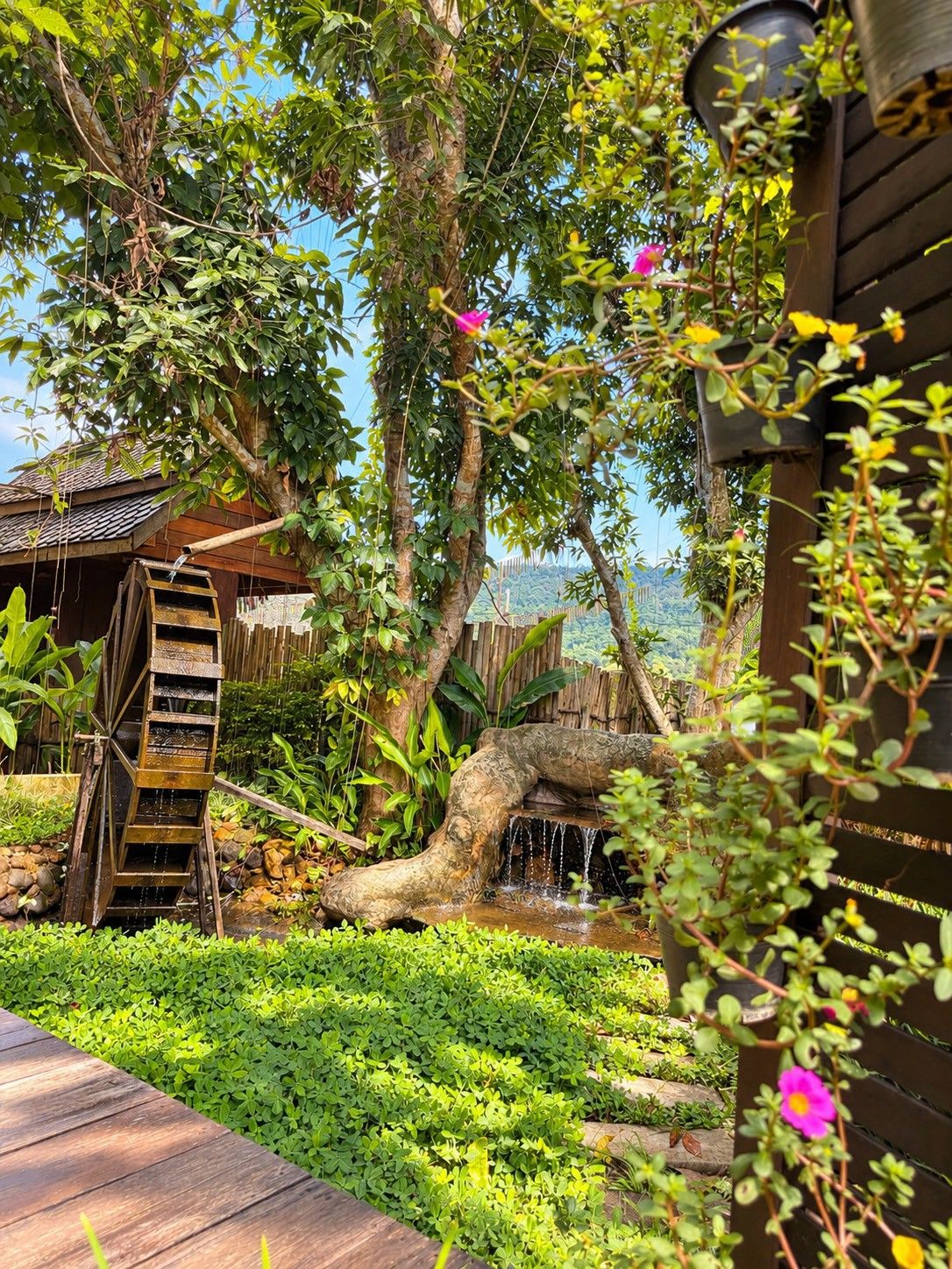 A traditional water wheel, lush greenery, and a wooden structure with a thatched roof.