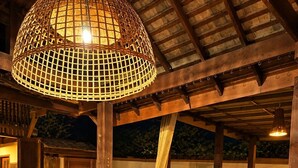 Uma área de refeições ao ar livre com uma mesa de madeira redonda e cadeiras de vime, sob uma pérgola de madeira com luminária trançada.